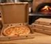 Boîtes à pizza en carton kraft de différents formats avec une pizza ouverte devant le four d’une pizzeria professionnelle.